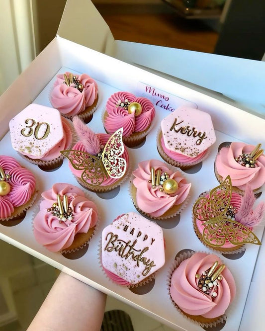 Box of pink cupcakes with gold decorations, including numbers and 'Happy Birthday' text, held by a person.